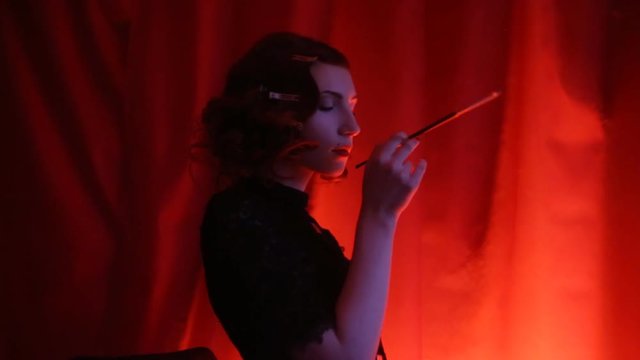 Redhead woman with curly hair in a black dress and retro makeup with pen on a red background. Red-haired girl with fatal face in red room. Noir woman silhouette smoking a cigarette in the mouthpiece 