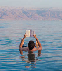 Young man reads a book floating in the waters of the Dead Sea in Israel