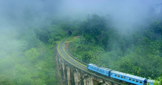 Train crossing Nine Arches Bridge and enters tunnel in foggy morning with clouds of mist above mountains overgrown with green tropical forest in Ella Sri Lanka