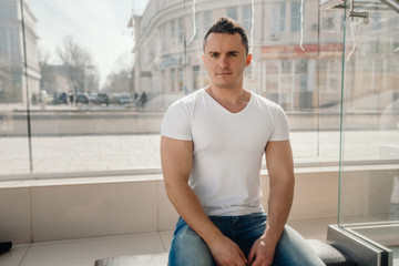 The man walks down the street in one of sunny days. He has come into shopping center and has decided to sit down to have a rest. He pleasant looks. On him a white t-shirt.