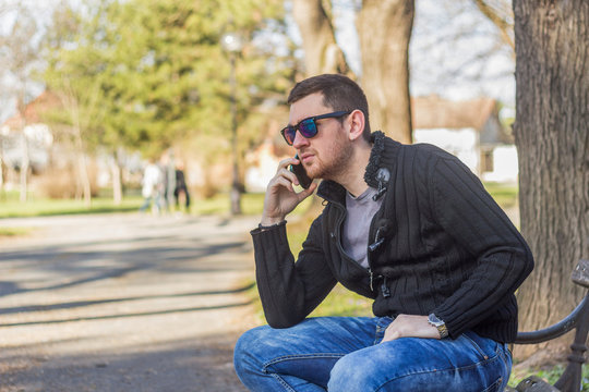 Handsome Man Talking On Phone While Sitting On Bench In Park