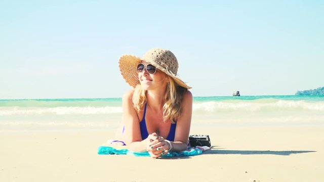 junge frau im blauen bikini liegt am strand mit einer vintage kamera und hat einen sonnenhut auf