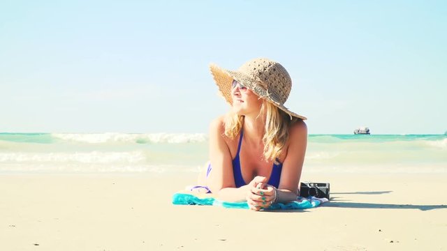 junge frau im blauen bikini liegt am strand mit einer vintage kamera und hat einen sonnenhut auf