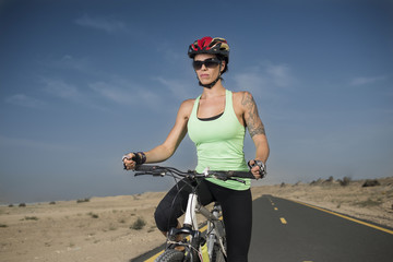 Closeup of beautiful woman wearing a bicycle helmet and sports glasses with tattoos riding a mountain bike on the road with a desert background 