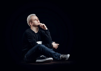 Pensive young man sitting on floor. Casual style. Black background. 