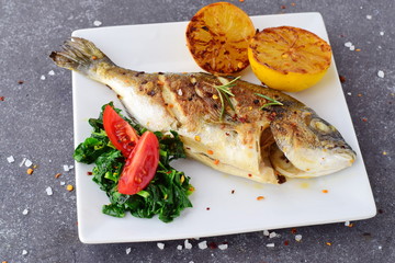 Grilled dorado with grilled lemov, spinach and tomato on a white plate on a grey abstract background. Healthy eating concept. Mediteranean lifestyle
