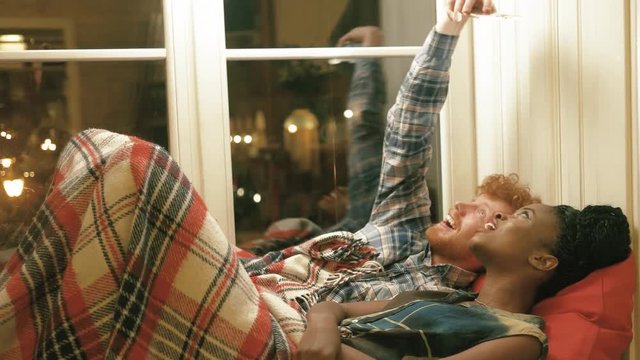 The man with golden hair and beard and his afro-american girlfriend are taking selfies, making funny faces while lying on the window-sill under the red knitted plaid ner the window. Lovely couple