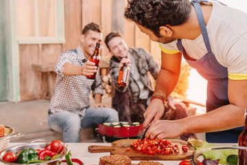 man making burgers while his friends drinking beer sitting behind on porch