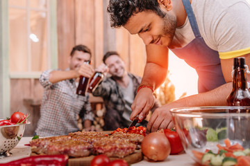 man making burgers while his friends drinking beer sitting behind on porch
