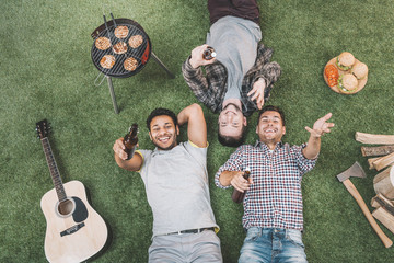 Top view of happy young men lying on grass with beer bottles while grilling meat at picnic