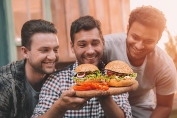 Three smiling men looking at fresh homemade hamburgers on wooden board