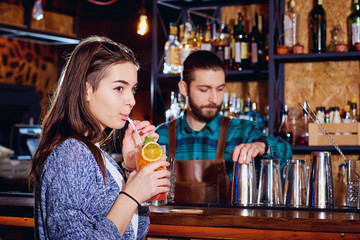 Girl drinks a cocktail from straw in bar on barman's background.