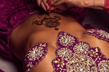 White woman a mehendi artist paints an ornament of henna on an eastern beautiful girl’s stomach