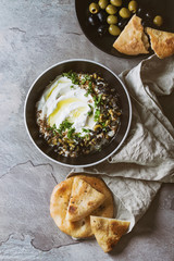 labneh middle eastern lebanese cream cheese dip with olive oil, salt, herbs, olives tapenade served in black bowl with traditional pita bread over gray texture metal background. Top view with space