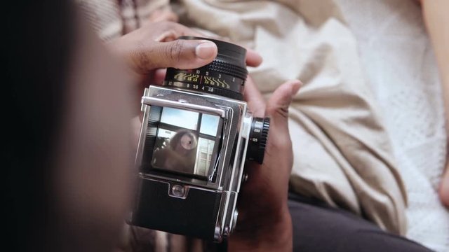 Close-up view of african man holding old photocamera, taking photo of woman near window. Multiracial couple on the bed.