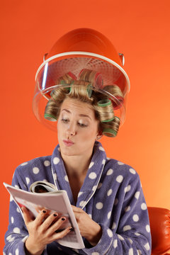 Eine Frau mit Lockenwickler für Dauerwelle unter Trockenhaube sitzend liest eine Illustrierte