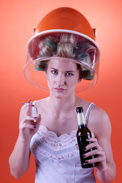 Eine Frau in Unterwäsche sitzt mit Zigarette und Bierflasche in der Hand unter Frisierhaube