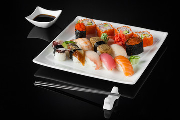 sushi in a white plate with sauce pot and chopsticks on a black background with reflection 