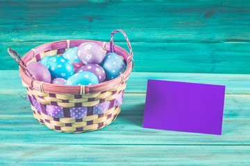 Violet and blue Easter eggs in a basket with a card. Blue wooden background. Happy easter.