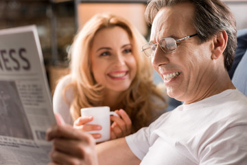 Happy middle aged couple sitting on bed, man reading newspaper at home