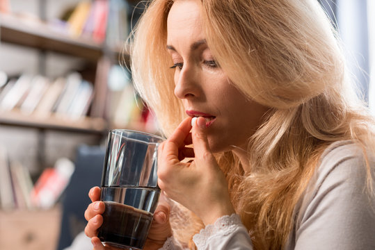 Blonde middle aged woman holding glass with water and taking pill