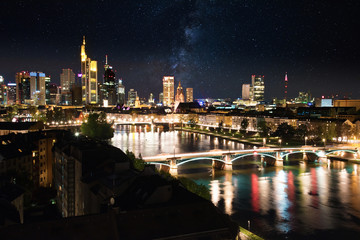 Fototapeta na wymiar illuminated skyline of Frankfurt am Main downtown financial district seen from the east with river in foreground at night with sky full of stars