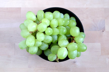 Lot of ripe green grape berries on bunche in black round bowl on wooden background top view close up