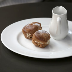 A jug with hot chocolate and two eclairs with cream on a plate on a table in a cafe