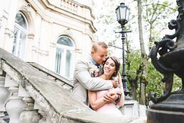 Blond groom hugs bride from behind standing with her on the stairs to old European house