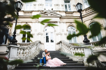 Look from behind green bushes at wedding couple sitting on the stone stairs to an old house
