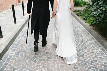 Look from behind at groom in tailcoat and bride in dress with long train walking on the road