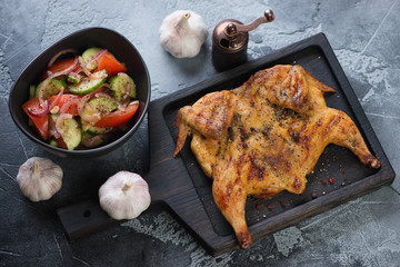 Wooden serving board with roasted chicken tabaka and vegetable salad on a grey stone background