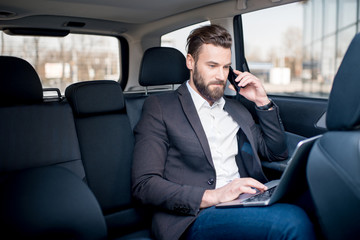 Handsome businessman talking with phone sitting with laptop on the backseat of the car - obrazy, fototapety, plakaty