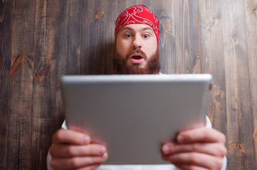 Shocking news online. Surprised young man with beard holding digital tablet against wooden wall.