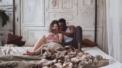 Multiracial couple in pajamas sitting on bed and use smartphone. African man and european woman looking photos, laughing - Powered by Adobe