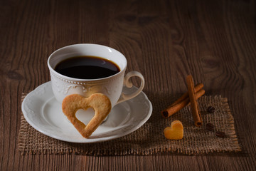 a cup of coffee and cookies like heart, cinnamon, coffee beans on wooden table on dark background