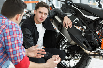 Tell me about this one. Portrait of a young handsome customer listening to motorcycle salon manager posing near a new motorbike