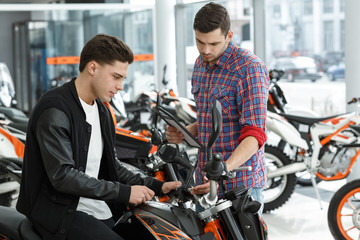 Quality guaranteed. Young male motorcycle salon manager showing his cheerful customer a new motorbike for sale