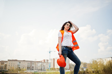 Engineer builder woman in uniform waistcoat and orange protective helmet against new buildings.