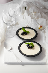 Delicious tasty Italian dessert Panna Cotta with fresh blueberries and mint leaves in black ramekins. Served with vine glasses, white tea spoon and orchid flowers for decoration.
