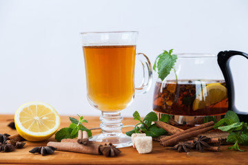 Clear big glass and teapot of hot black herbal tea with lemon, mint and Melissa leaves on light rustic wooden table. Summer, Autumn, winter drink. Cinnamon and cane sugar for decoration.
