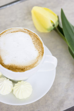 Coffee drink cappuccino in a white cup on a table in a cafe next to the yellow flowers of gorse and tulip