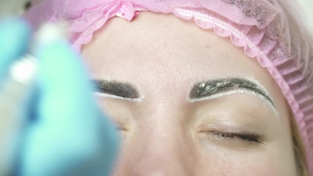 Cosmetologist is making an eyebrow permanent makeup in salon closeup