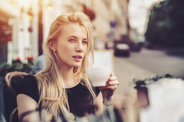 Adult blonde lady is drinking hot tea or coffee and thoughtfully looking aside from street cafe while enjoying her leisure time alone, portrait of beautiful woman lunching in cafe during work break
