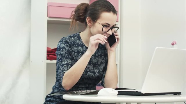Young attractive female fashion designer using computer laptop and talking on mobile phone call in manufacturing office studio