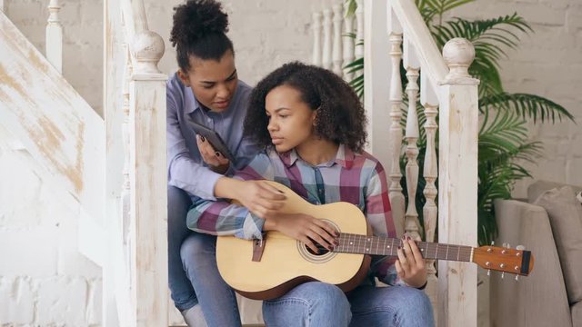 Mixed race young woman sitting on stairs teaching her teenage sister to play acoustic guitar at home