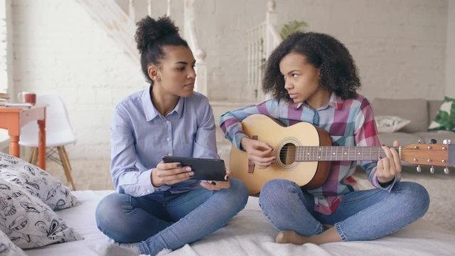 Mixed race young woman with tablet computer sitting on bed teaching her teenage sister to play acoustic guitar at home