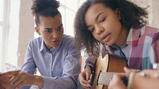 Mixed race young woman with tablet computer sitting on bed teaching her teenage sister to play acoustic guitar at home