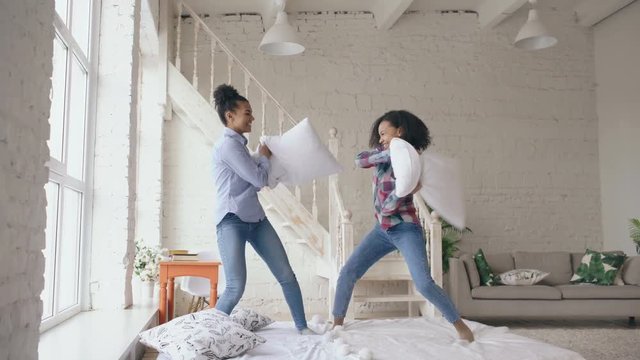Mixed race young pretty girls jumping on bed and fight pillows having fun at home