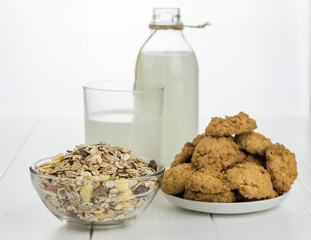 Milk bottle, bowl with muesli and fresh baked cookies on a white wooden table.
